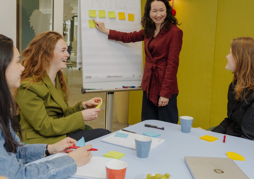 Lisa (meest links) zit, Serra (links) zit, Jill (midden) staat en wijst naar post-its op flip-over, Luna (rechts) zit